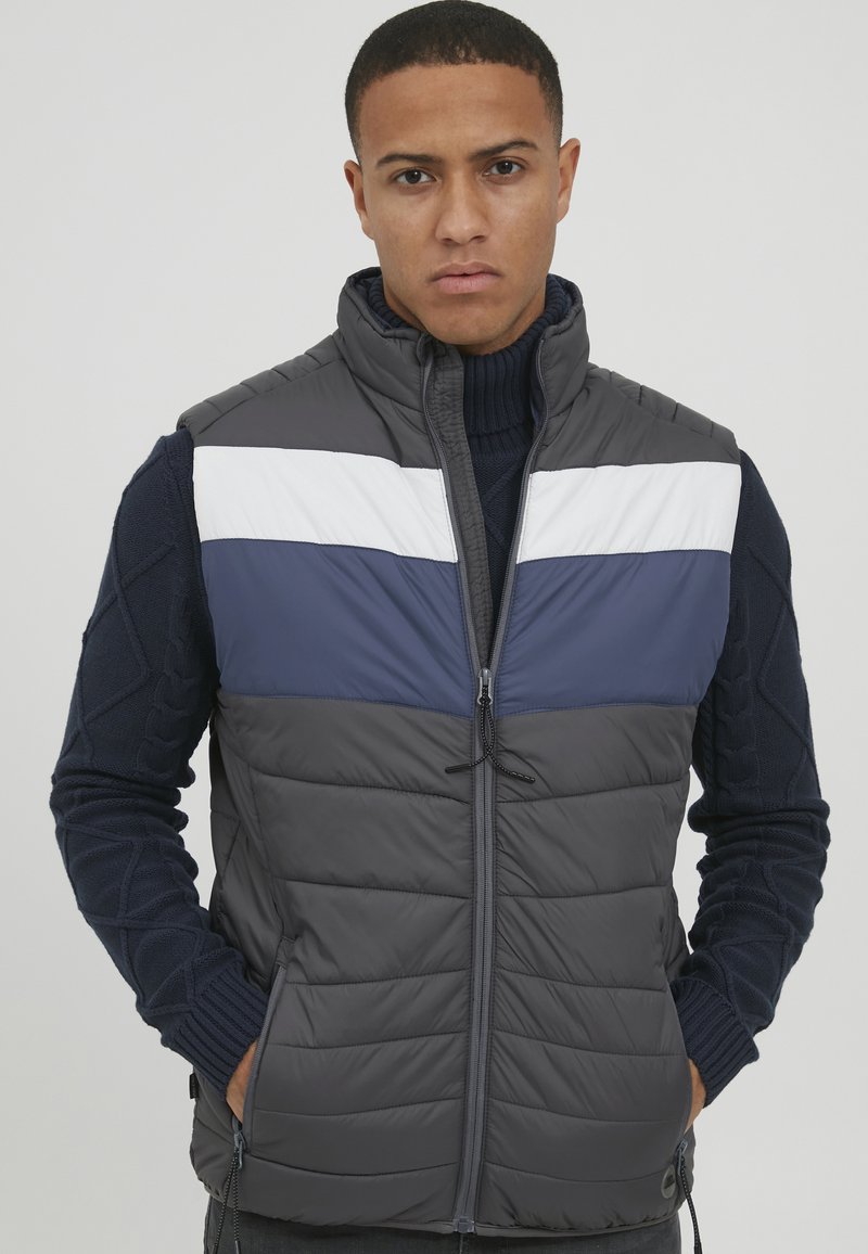 Vesta puffy gri cu dungi orizontale bleumarin și alb, guler înalt, fermoar frontal și tiv elastic; mâneci din tricot texturat.
