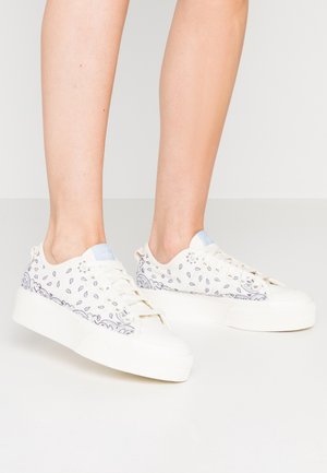 Des baskets en toile blanches présentent un accent bleu sur la languette, décorées de petits motifs en forme de goutte violette le long des côtés, et d'une épaisse semelle blanche.