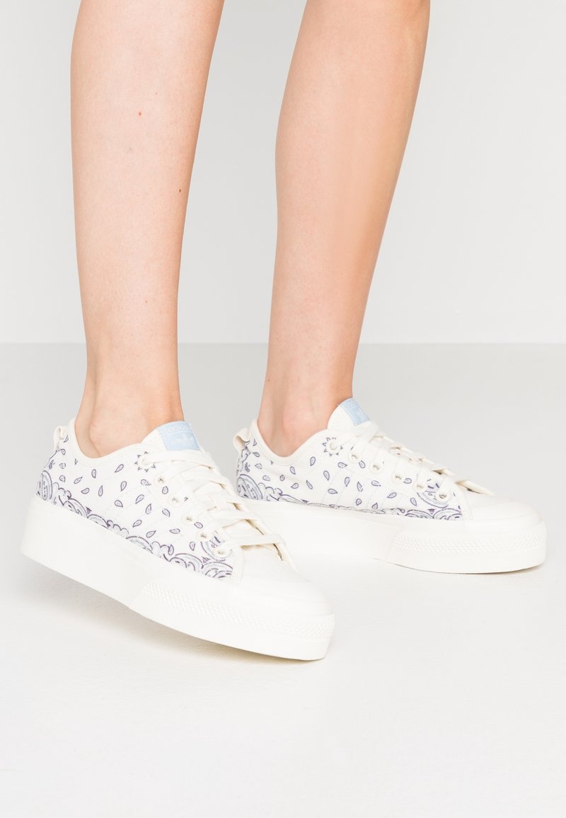 Vita kanvas sneakers med en blå accent på tungan, dekorerade med små lila tårformade mönster längs sidorna och en tjock vit sula.