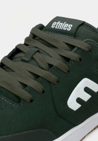 Πράσινα σουέτ παπούτσια σκεϊτ με επίπεδα κορδόνια, διαθέτουν λευκό λογότυπο στο πλάι, υφή επιφάνειας και ελαστική σόλα.