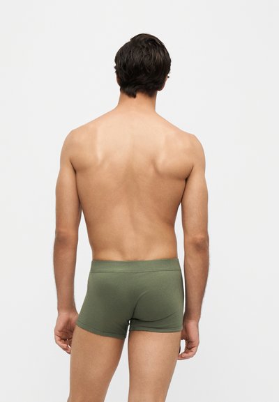 Homme debout de dos, portant un boxer vert sur un fond gris clair uni.