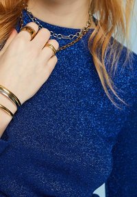 Haut bleu scintillant avec une surface texturée. Les accessoires comprennent une chaîne argentée à plusieurs rangs, plusieurs bagues ainsi que trois bracelets métalliques.