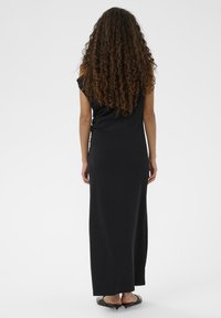 Kvinde med langt krøllet hår iført en sort ærmeløs maxikjole og sorte spidse lavhælede sko, stående foran en ensfarvet hvid baggrund.