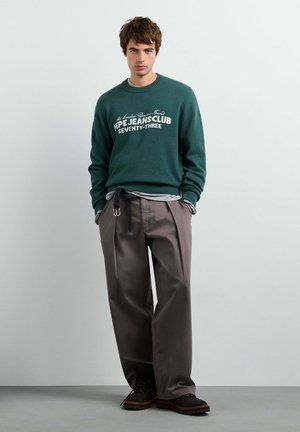 Giovane uomo che indossa un maglione verde con testo bianco e pantaloni grigi larghi, in piedi davanti a uno sfondo grigio chiaro uniforme.