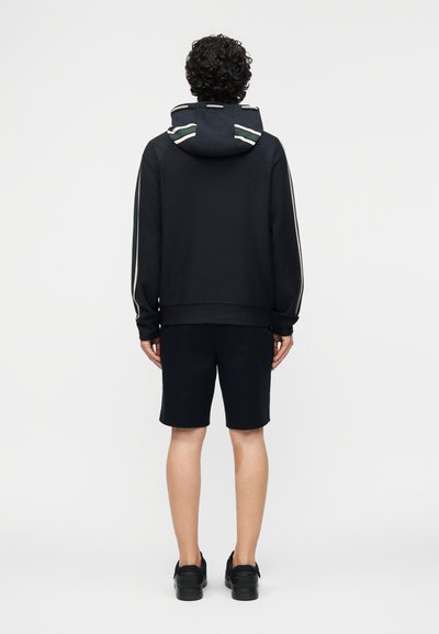 Άνδρας που στέκεται με την πλάτη, φορώντας μαύρο φούτερ με ρίγες στη κουκούλα και στα μανίκια, μαύρο σορτς και μαύρα αθλητικά παπούτσια.