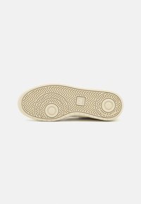 Krämfärgad skosula med fiskbensmönster, utrustad med cirkulära grepppunkter och en upphöjd logotyp. Slät gummimaterial.