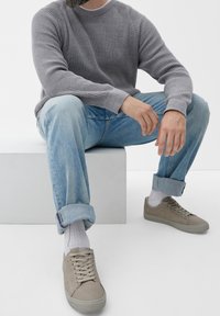 Mann mit Bart, der hellblaue hochgekrempelte Jeans, einen grauen Rippstrickpullover, weiße Socken und beigem Sneakern trägt und auf einem weißen Block sitzt.
