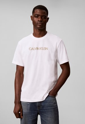 Άνδρας που φοράει λευκό t-shirt Calvin Klein με μπεζ λογότυπο και μπλε τζιν, στέκεται με το ένα χέρι στην τσέπη μπροστά σε ανοιχτό γκρι φόντο.