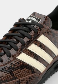 Sneaker in pelle di serpente marrone con lacci neri e tre strisce bianche, caratterizzati da una finitura testurizzata e una suola in gomma nera testurizzata.