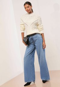 Weißer Pullover mit Zopfmuster und hohem Kragen, kombiniert mit hellblauen, weitem Denim-Jeans. Das Modell hält eine schwarze Clutch und trägt schwarze Absatzschuhe.