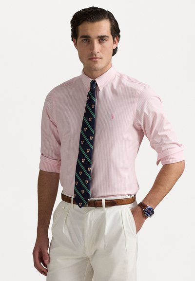 Chemise à rayures rose clair avec col boutonné, assortie d'une cravate bleu marine ornée de motifs d'emblèmes colorés et de pantalons beige foncé.