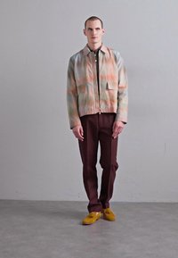 Giacca chiara a fantasia con due tasche frontali, abbinata a pantaloni borgogna scuri. Footwear giallo, con una texture liscia e un design minimale.