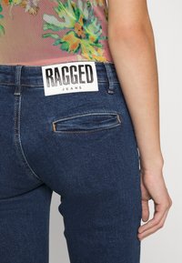 Närbild av en person som bär mörkblå jeans med en "RAGGED JEANS"-etikett på bakre midjan och en färgglad blommig topp.