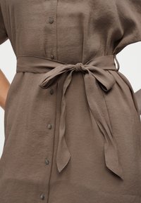 Καφέ φόρεμα με κουμπιά και υφασμάτινη ζώνη δεμένη σε φιόγκο στη μέση, που παρουσιάζει υφή και ελαφριές ρυτίδες στο υλικό.