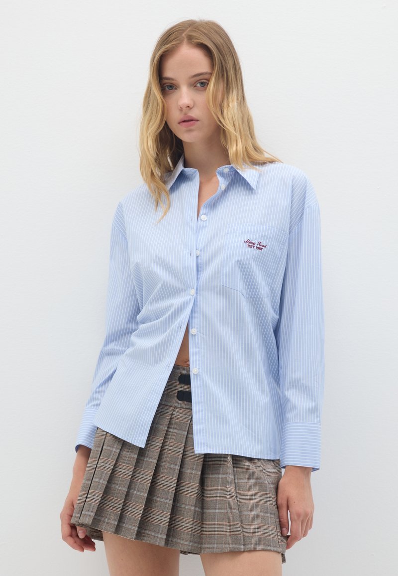 Camicia azzurra chiara a righe con taschino e logo ricamato; abbinata a una gonna pieghettata a quadri marrone. Tessuto morbido e liscio.