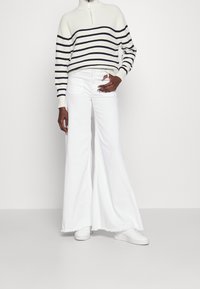 Jeans bianchi a campana abbinati a un maglione a righe crema con colletto alto e righe blu navy, che presenta una texture a costine e maniche cuffiate.