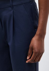 Δεξί χέρι ενός ατόμου με σκούρο χρώμα δέρματος που ακουμπάει δίπλα σε σκούρο μπλε παντελόνι με ορατή μπροστινή τσέπη και θηλιά ζώνης.