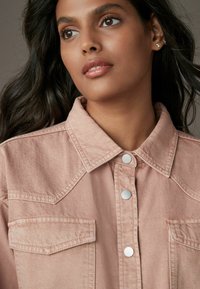 Giacca di jeans rosa chiaro con colletto strutturato, bottoni a scatto in metallo e due tasche sul petto. Il tessuto ha una leggera texture.