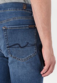 Jean en denim bleu pour hommes avec une poche arrière dotée d'un motif de couture sombre, des accents de couture orange et une patch de la marque sur la taille.