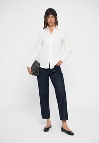 Camicia bianca con colletto a punta, abbinata a jeans scuri a sigaretta e ballerine nere, mentre tiene una borsa nera texture.