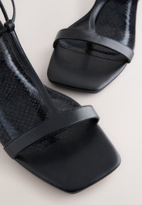 Sandales noires à bout ouvert avec avant carré, bride unique sur les orteils et semelle intérieure texturée rappelant un motif peau de serpent.