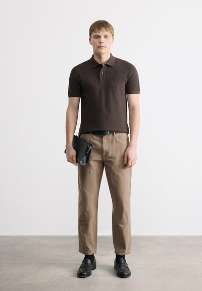 Jeune homme debout, portant un polo marron foncé, un pantalon beige, une ceinture et des chaussures noires, tenant un porte-documents en cuir noir dans une main.