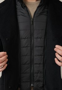 Czarny wełniany płaszcz na czarnej kurtce puchowej, z zamkiem błyskawicznym z przodu i pikowanym wzorem. Pod spodem widoczny beżowy sweter.