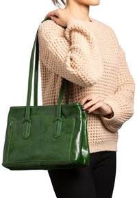 Borsa in pelle verde con due manici lunghi, superficie liscia, forma rettangolare e cuciture visibili. Indossata da una persona in un maglione beige.