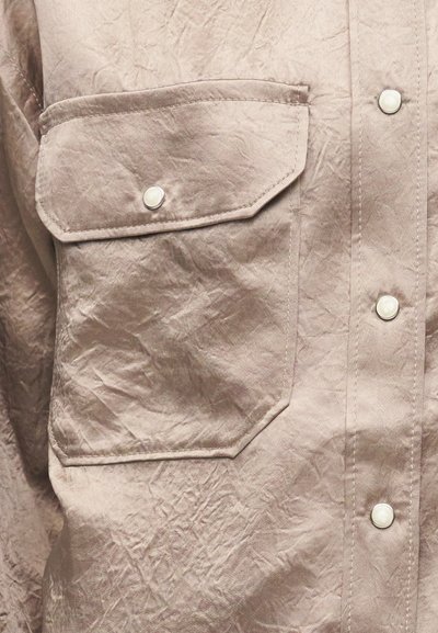 Μπεζ πουκάμισο από ζαρωμένο ύφασμα με μεγάλη μπροστινή τσέπη με καπάκι και κλείσιμο με κουμπί-σούστα, καθώς και επιπλέον κουμπιά-σούστες στη μπροστινή πλευρά.