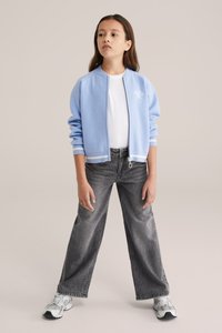 Svetlo modra bomber jakna z zadrgo in belimi poudarki, oblečena čez belo majico. Kombinirana z ohlapnimi, zbledelimi črnimi kavbojkami in športnimi čevlji.