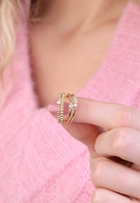 Set van drie gouden ringen, met één ring met kralen, een andere met gladde banden, en de derde versierd met kleine heldere stenen, vastgehouden tussen de vingers.