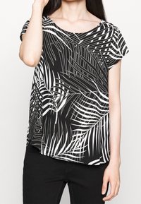 Donna che indossa un top a maniche corte bianco e nero con un motivo astratto di foglie di palma e pantaloni neri, in posa con una mano vicino al viso.