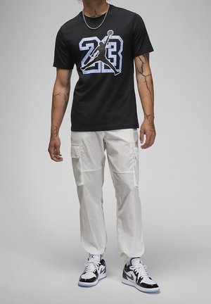 Mężczyzna ubrany w czarną koszulkę Jordan z numerem "23" i logo Jumpman, białe spodnie cargo oraz czarno-białe buty sportowe Nike, stojący na szarym tle.