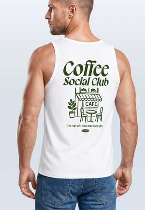 Mann trägt weißes Tanktop mit grünem "Coffee Social Club"-Text und Café-Illustration auf dem Rücken, kombiniert mit blauer Jeans.