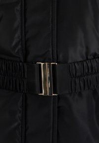 Mustapuffatakkia, jossa on teksturoitu pinta, pleattyjä yksityiskohtia ja kultasävyinen vyösolki vyötäröllä säätämistä varten.