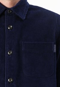 Camisa de pana azul marino con cierre de botones en la parte delantera, cuello puntiagudo y un bolsillo en el pecho. Presenta rayas verticales texturizadas y botones negros.