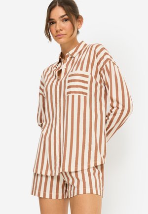 Frau trägt braun-weiß gestreiftes Langarmshirt und passende Shorts, steht mit den Händen hinter dem Rücken vor weißem Hintergrund.