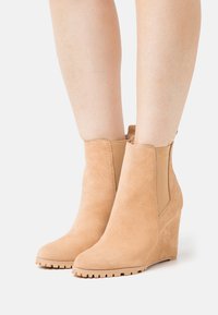 Botas de tornozelo em camurça bege com salto tipo cunha e painéis laterais elásticos, apresentando uma sola de borracha texturizada e um design de bico arredondado.