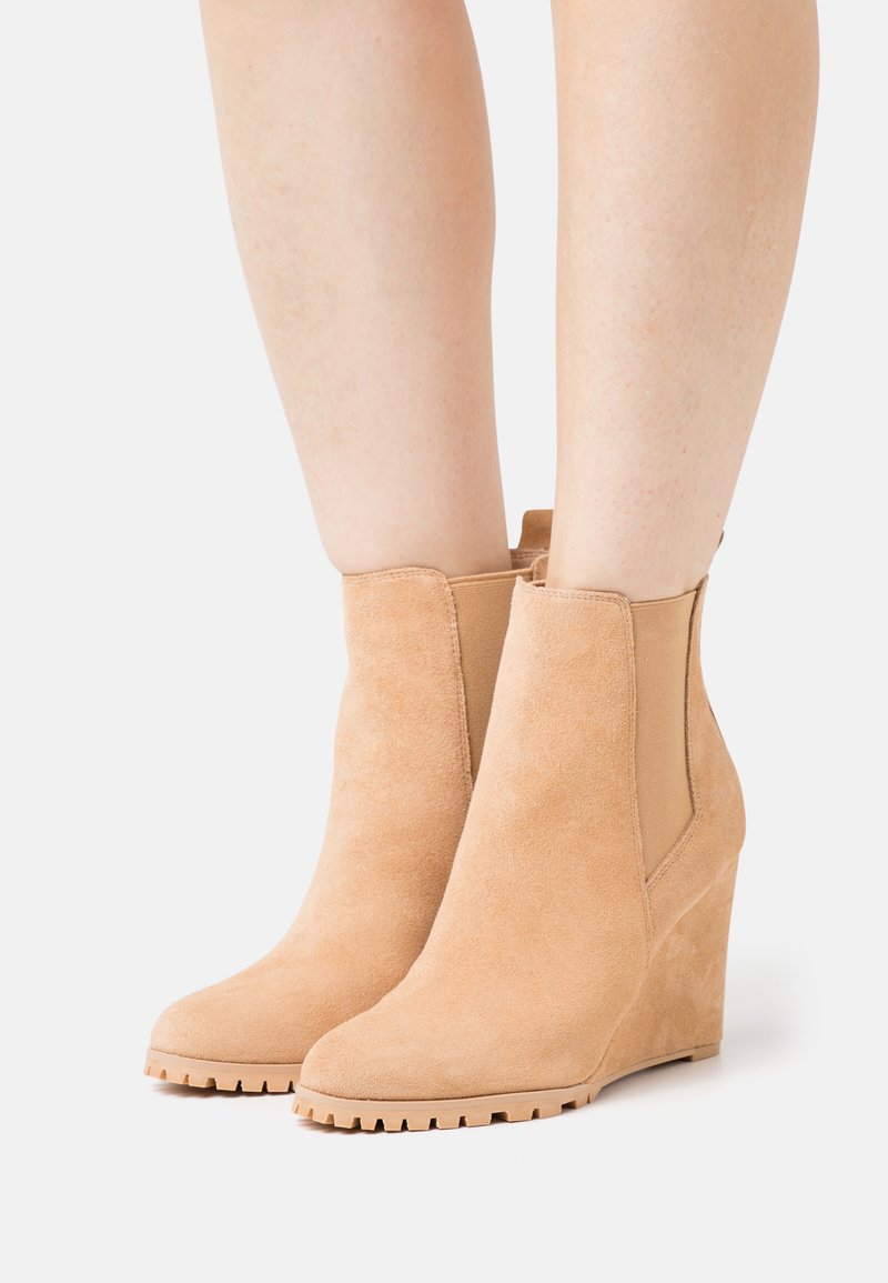 Botas de tornozelo em camurça bege com salto tipo cunha e painéis laterais elásticos, apresentando uma sola de borracha texturizada e um design de bico arredondado.