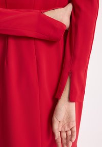 Czerwona sukienka z długimi rękawami, z boku zapinana na suwak, uszyta z gładkiego, płynnego materiału. Końce rękawów mają małe rozcięcia, co ułatwia ruch.