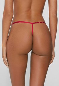 Červené tanga s tenkými pásky a malými kovovými doplňky, představující minimalistický design a hladkou texturu.