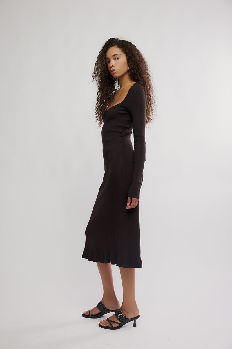 Vestido preto de malha canelada com mangas compridas, decote redondo e bainha evasê. Combinado com saltos pretos de tiras. O cabelo é encaracolado.