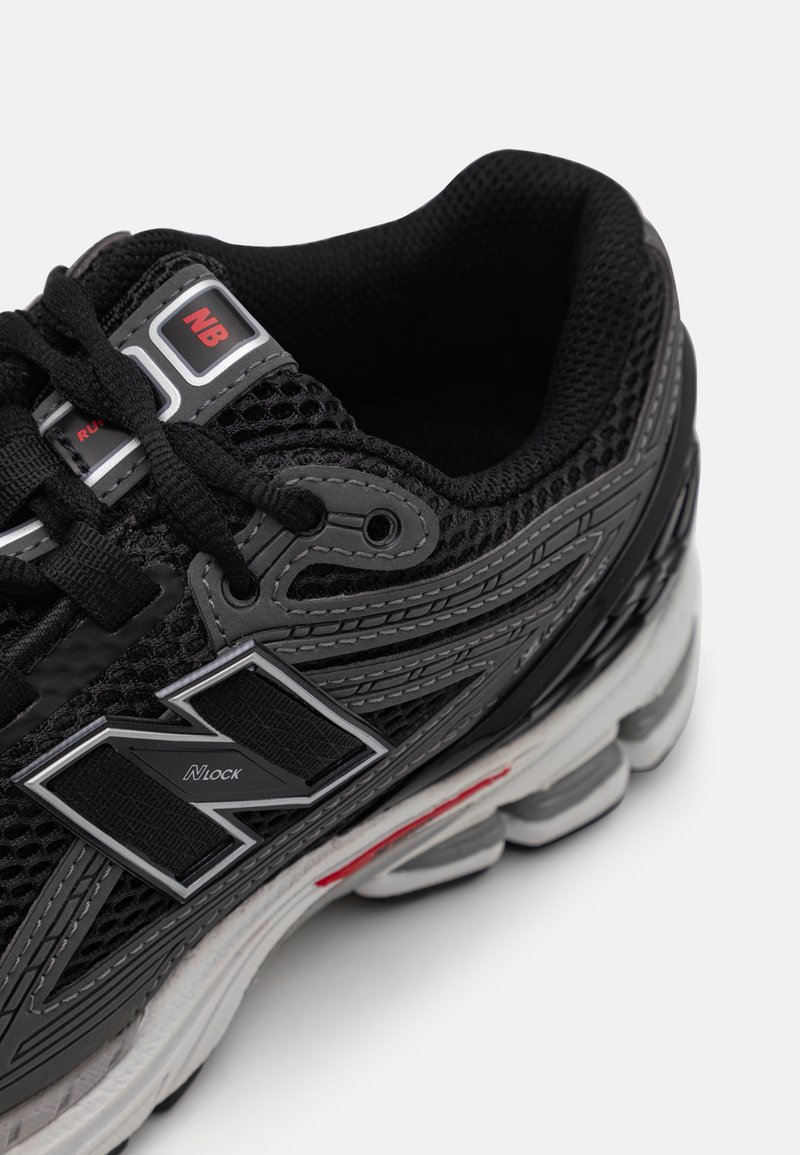 Zapato atlético negro con parte superior de malla, con acentos grises y un prominente logo N. Suela gruesa blanca y gris con detalles de color rojo.