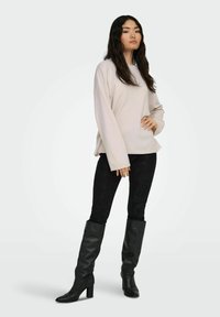 Top de manga larga color beige claro con un ajuste relajado, combinado con pantalones negros texturizados y botas de cuero negras hasta la rodilla con un tacón cuadrado.