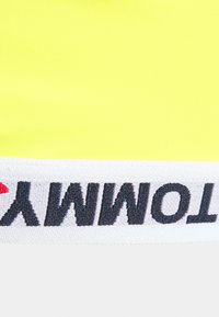 Top desportivo amarelo cropped com um tecido suave e elástico e uma faixa elástica branca na parte inferior com "TOMMY" em letras pretas.