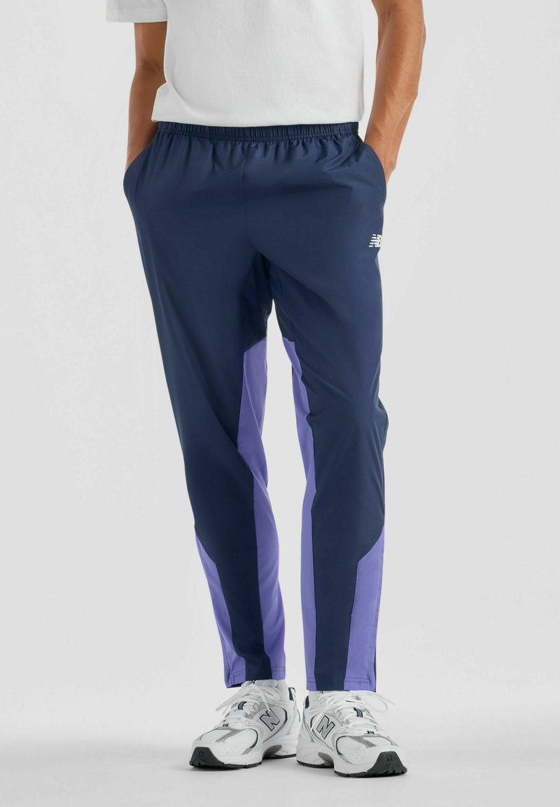 Person wearing navy blue and purple track pants with white New Balance sneakers, standing with hands in pockets against a plain background.
