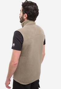 Mann mit dunklem Haar und Bart, der eine beige Fleeceweste über einem schwarzen kurzärmligen Hemd trägt und mit dem Rücken zur Kamera steht.