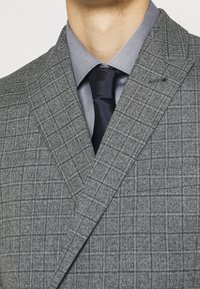 Giacca da completo su misura grigia a quadri con revers a incavo, abbinata a una camicia azzurro chiaro e cravatta scura, che mette in risalto un tessuto lavorato.