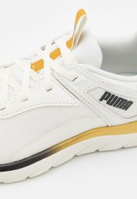 Bílé tenisky s síťovaným svrškem, žlutým akcentem u šňůrek a jemným zlatým přechodem na podrážce. Obsahují černé detaily a logo.