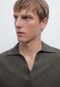 Tmavě olivově zelené textured polo tričko s výstřihem do V a krátkým límečkem, nošené osobou s krátkými vlasy a neutrálním výrazem.